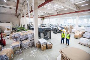 Workers of paper recycling factory wearing reflective vests standing in shop with packaged papers in cartoon and cutting machines